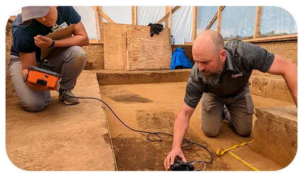 Dos técnicos trabajan en una excavación arqueológica, utilizando un georradar para la arqueología y estudios forenses. Uno de ellos sostiene el dispositivo y lo posiciona cuidadosamente sobre el suelo, mientras que el otro monitorea y analiza los datos en una pantalla. El entorno muestra un área de excavación delimitada y organizada, con herramientas y materiales de trabajo a su alrededor, lo que sugiere una investigación meticulosa y detallada.
