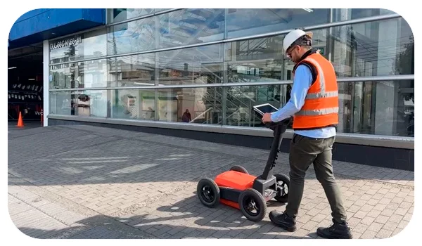 Un técnico con casco y chaleco de seguridad opera un georradar en una acera, frente a un edificio con grandes ventanales. El dispositivo se utiliza para la identificación de tuberías y servicios públicos subterráneos, asegurando que se puedan realizar excavaciones y trabajos de mantenimiento de manera segura y eficiente. El entorno urbano sugiere que esta operación es parte de un proyecto de infraestructura en una zona comercial o residencial.