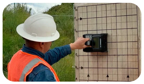 Un técnico, con casco y chaleco de seguridad, opera un radar de concreto contra una pared con una cuadrícula marcada. Este dispositivo se utiliza para detectar estructuras internas y posibles fallas en la pared, asegurando la integridad del material y facilitando las tareas de inspección y mantenimiento. El entorno muestra vegetación en el fondo, indicando que la operación se realiza en una zona al aire libre.