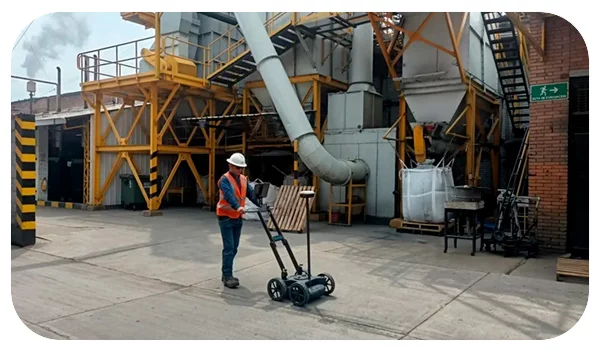Un técnico realiza una inspección en una planta industrial utilizando un georradar (GPR) para la detección de tuberías de aceite y gas. El técnico, equipado con casco y chaleco de seguridad, maneja el georradar sobre una superficie de concreto, rodeado de estructuras industriales y maquinaria pesada.